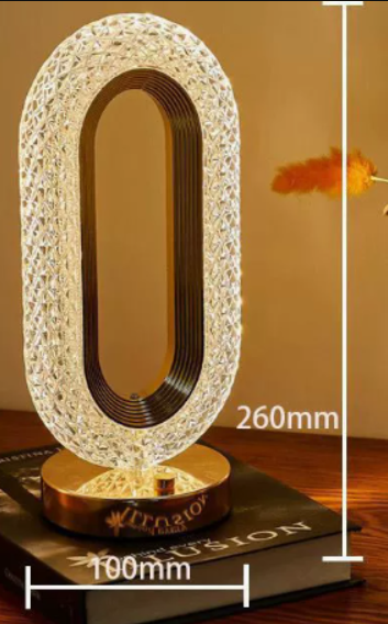 A modern oval-shaped table lamp with a textured, faceted clear crystal-like frame and a gold-toned circular base, shown on a wooden surface with overlaid dimension lines indicating 260mm height and 100mm width.